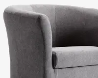 Nærbillede af en moderne lænestol betrukket med tekstureret gråt stof. Stolen har en buet ryg og armlæn, der flyder sammen i et elegant design og giver et blødt og komfortabelt udtryk. Sædepuden er tyk og blød, hvilket giver god støtte. Syningerne er diskrete og pæne, hvilket understreger stolens rene sop moderne udseende.