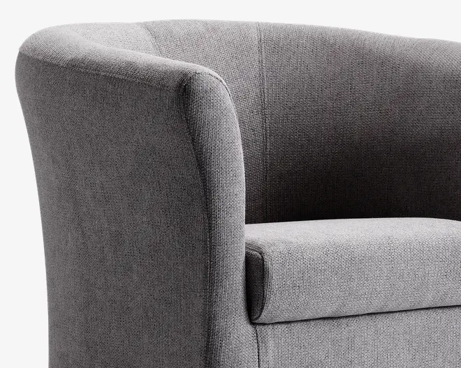 Nærbillede af en moderne lænestol betrukket med tekstureret gråt stof. Stolen har en buet ryg og armlæn, der flyder sammen i et elegant design og giver et blødt og komfortabelt udtryk. Sædepuden er tyk og blød, hvilket giver god støtte. Syningerne er diskrete og pæne, hvilket understreger stolens rene sop moderne udseende.