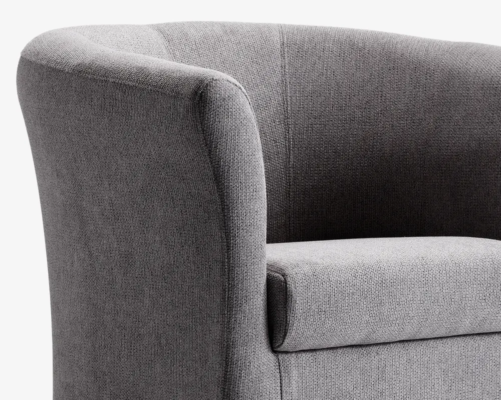 Nærbillede af en moderne lænestol betrukket med tekstureret gråt stof. Stolen har en buet ryg og armlæn, der flyder sammen i et elegant design og giver et blødt og komfortabelt udtryk. Sædepuden er tyk og blød, hvilket giver god støtte. Syningerne er diskrete og pæne, hvilket understreger stolens rene sop moderne udseende.