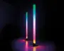 To lodrette LED-lysstave med et slankt, moderne design står på flade baser. Hver stav udsender en farverig gradient med nuancer af grøn, blå, pink og gul, hvilket skaber en glødende effekt på overfladen nedenunder. Lysene er forbundet med synlige strømkabler, og belysningen er kraftig nok til at kaste farverige refleksioner på det omkringliggende område.