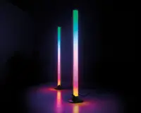 To lodrette LED-lysstave med et slankt, moderne design står på flade baser. Hver stav udsender en farverig gradient med nuancer af grøn, blå, pink og gul, hvilket skaber en glødende effekt på overfladen nedenunder. Lysene er forbundet med synlige strømkabler, og belysningen er kraftig nok til at kaste farverige refleksioner på det omkringliggende område.