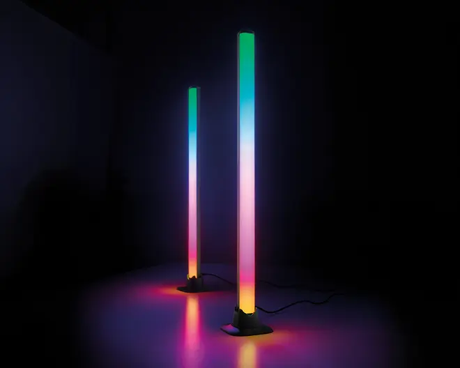 To lodrette LED-lysstave med et slankt, moderne design står på flade baser. Hver stav udsender en farverig gradient med nuancer af grøn, blå, pink og gul, hvilket skaber en glødende effekt på overfladen nedenunder. Lysene er forbundet med synlige strømkabler, og belysningen er kraftig nok til at kaste farverige refleksioner på det omkringliggende område.