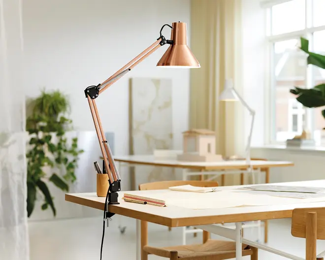 Justerbar skrivebordslampe med kobberfarvet metalskærm og arm, sorte metalled og en klemmefod til fastgørelse på bordkanten. Lampen har et moderne, industrielt design og en fleksibel arm, der gør det nemt at justere lysets placering. Strømkablet er synligt og hænger ned fra lampens base.