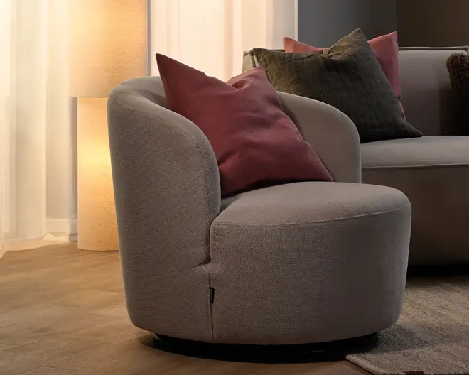Rund polstret loungestol i lysegrå taupe-stof med lavt, bredt sæde, buet ryg og armlæn samt sort rund base. Stylet med en bordeaux pyntepude.