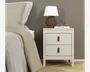 Et moderne natbord med mat, lysebeige finish og to rummelige skuffer. Hver skuffe har et unikt greb lavet af brunt læder med en messingknap. Natbordet står på fire lige ben, hvilket giver en let forhøjning fra gulvet. Toppen er dekoreret med en geometrisk brun lampe med hvid skærm, et klart glas og en hvid bog med titlen 'SCULPTURE'. Designet er minimalistisk og funktionelt, velegnet til moderne soveværelser.