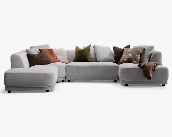 En stor, moderne hjørnesofa betrukket med lysegråt stof. Sofaen har brede, bløde siddepladser og lave, afrundede armlæn. Den er pyntet med forskellige pyntepuder i jordfarver, herunder brune, rustfarvede, olivengrønne og beige nuancer samt en brun pude med mønster. Et blødt, brunt tæppe er lagt over det ene armlæn, hvilket giver et hyggeligt udtryk. Sofaens design er moderne og indbydende, velegnet til en rummelig stue.