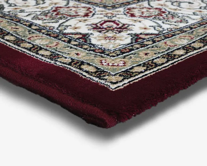 Nærbillede af et tykt, blødt tæppehjørne med en dyb bordeaux kant og et detaljeret, ornamenteret mønster med blomster i creme, beige og røde nuancer. Tæppet har en blød, tæt luv og en veldefineret kant, hvilket vidner om god kvalitet og komfort.