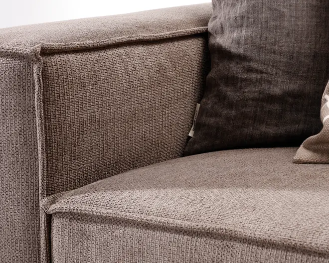 Nærbillede af et sofahjørne med tekstureret, lysebrunt betræk og synlige syninger langs kanterne. Sædet og armlænet har et firkantet, moderne design. En mørkebrun pude med diskret vævet mønster ligger op ad ryglænet og giver kontrast og komfort.