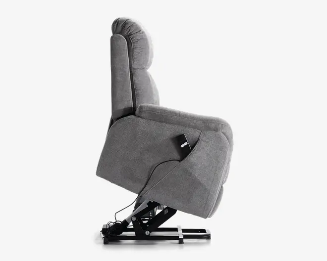 Sidebillede af en grå polstret lænestol med løftemekanisme. Stolen har en polstret ryg og armlæn for ekstra komfort. En ledningsforbundet fjernbetjening er fastgjort på siden, så stolen nemt kan justeres. Basen har en solid metalramme, der understøtter løfte- og tilbagelæningsfunktionerne.