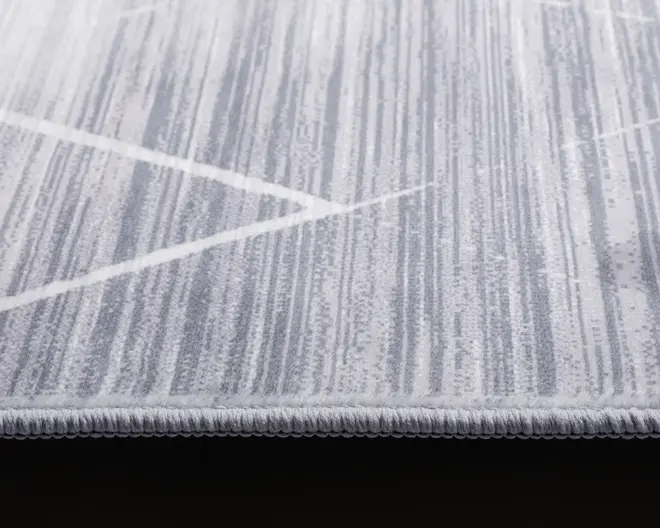 Nærbillede af et tæppe med moderne design, der har lodrette striber i forskellige grå og hvide nuancer. Tæppet har lav luv og en pænt afsluttet, tæt kantet kant for holdbarhed. Mønstret inkluderer diskrete hvide diagonale linjer, der krydser de grå striber og giver et moderne udtryk.