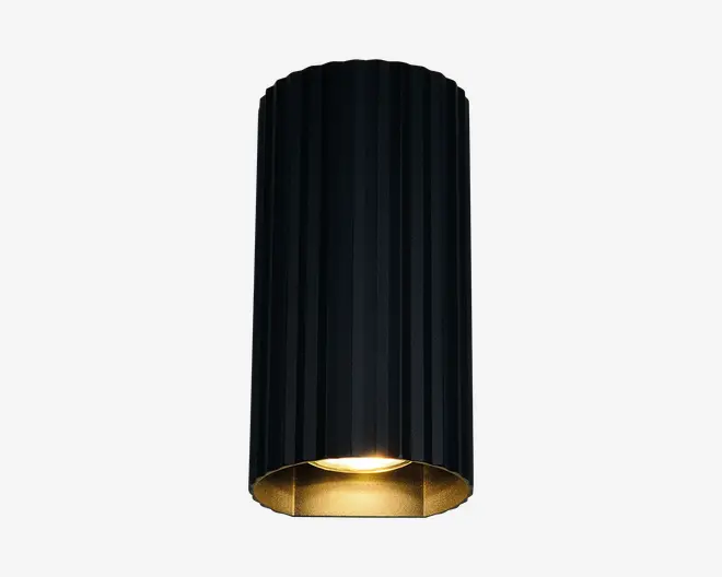 En cylindrisk lampe med lodrette riller og mat sort yderside samt en guldfarvet inderside. Lampen er åben i bunden, hvilket giver et direkte nedadgående lys. Det elegante og moderne design gør den velegnet til nutidige indretninger.