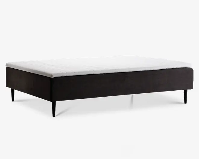 Minimalistisk sengebund med mørkt stofbetrukket ramme og slanke, koniske sorte ben. Toppen har en hvid, quiltet madraspude for ekstra komfort. Designet er moderne og enkelt, velegnet til forskellige soveværelsesstile.
