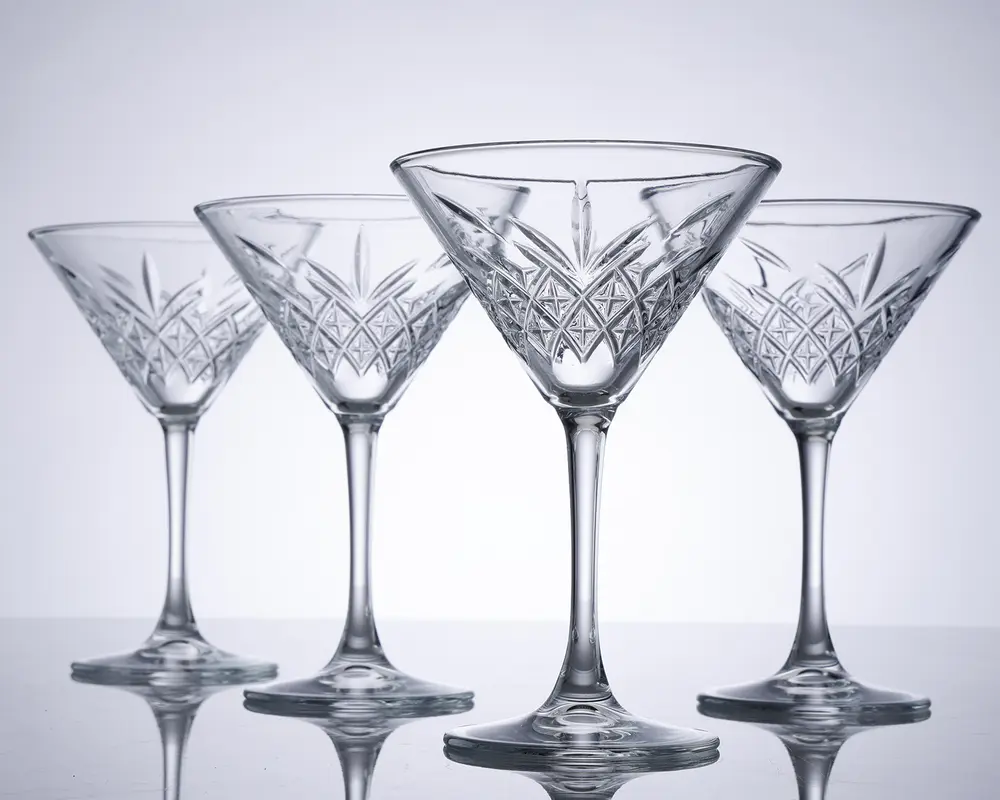 Sæt med fire klare krystalglas med bred, lav skål og lang stilk. Hvert glas har et indviklet slebet mønster med geometriske og vifteformede detaljer omkring skålen, hvilket giver dem et elegant og klassisk udseende. Glassene har en solid fod og er arrangeret på række, så det detaljerede håndværk fremhæves.