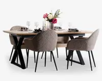 Et moderne spisesæt bestående af et rektangulært bord med lys træbordplade og sorte metalben i X-form. Sættet inkluderer seks polstrede stole i blødt, tekstureret beige stof med quiltet mønster på ryglænet og slanke sorte metalben. Bordet er stilfuldt dækket med hvide tallerkener, sorte servietter, klare vinglas og en blomsterdekoration i en glasvase som midtpunkt.