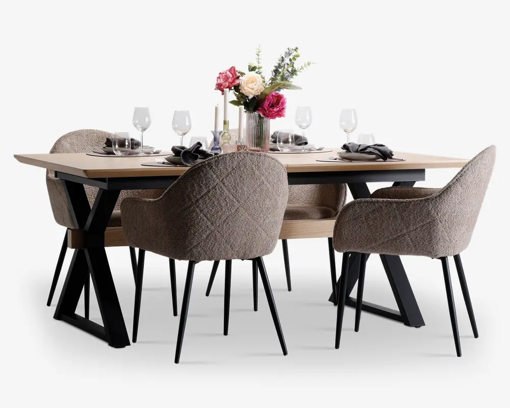 Et moderne spisesæt bestående af et rektangulært bord med lys træbordplade og sorte metalben i X-form. Sættet inkluderer seks polstrede stole i blødt, tekstureret beige stof med quiltet mønster på ryglænet og slanke sorte metalben. Bordet er stilfuldt dækket med hvide tallerkener, sorte servietter, klare vinglas og en blomsterdekoration i en glasvase som midtpunkt.
