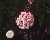 En træskål fyldt med individuelt indpakkede lyserøde og hvide skumfiduser formet som julemanden. Skålen står på en mørk overflade dekoreret med grangrene, kogler, et tændt hvidt stearinlys og rød-hvid snoet snor, hvilket skaber en festlig julestemning.