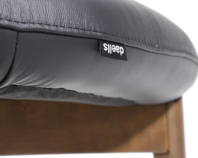 Nærbillede af en sædepude betrukket med sort læder og synlige syninger. Puden har et lille sort stofmærke med hvid tekst, fastgjort ved sømmen. Sædet hviler på en træramme med en glat finish.