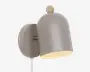 Væghængt lampe med mat taupefarvet kuppelformet skærm, rund vægplade og lille træfarvet knop på toppen, vist vinklet nedad med en varm lys inderside.