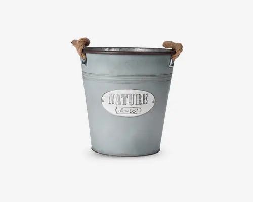 En rund metalbeholder med mat grå finish og to kraftige rebhåndtag fastgjort på hver side. Foran er der en oval metalplade med teksten 'NATURE' i store bogstaver og 'Since 1991' i mindre skrift nedenunder. Spanden er let konisk, bredere foroven og smallere forneden, hvilket giver den et rustikt og vintage udseende.