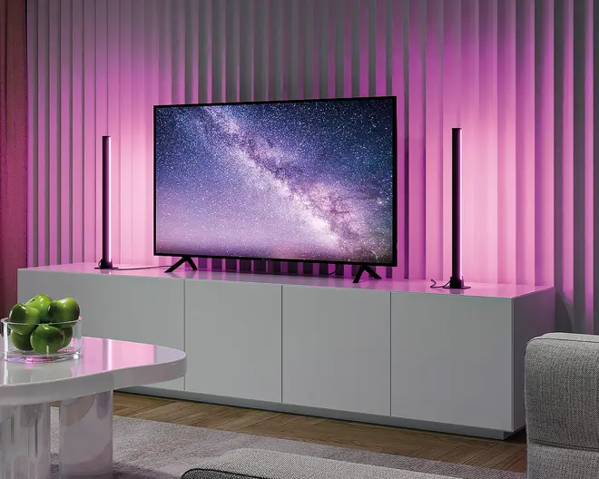 Moderne underholdningsopsætning med et fladskærms-tv, der viser en stjernehimmel, placeret på et elegant hvidt skab. To lodrette LED-lysstænger står på hver side af tv'et og udsender en blød pink glød, som oplyser den teksturerede væg bagved. Skabet har rene linjer og fire låger til opbevaring. I forgrunden står et rundt hvidt bord med en glas skål fyldt med grønne æbler, og en del af en lysegrå sofa ses til højre.