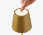 Nærbillede af en lampe i guldfarvet, mat finish med en berøringsfølsom tænd/sluk-knap på toppen. En hånd trykker på det oplyste tænd/sluk-symbol, hvilket viser, at lampen betjenes ved berøring. Lampen har et moderne, minimalistisk design med en let konisk cylindrisk form.