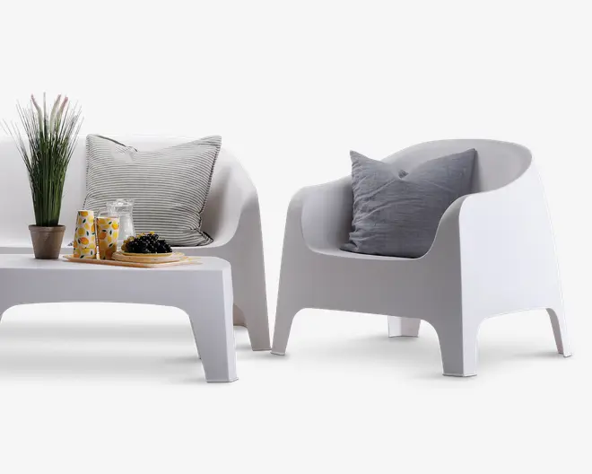 Moderne udendørs siddegruppe bestående af en hvid lænestol og en matchende to-personers sofa, begge med glatte, buede linjer og et minimalistisk design. Sættet inkluderer et lavt, rektangulært sofabord. Lænestolen og sofaen har hver en grå eller stribet pude for ekstra komfort. På bordet står en potteplante, en glaskande med citronskiver, to glas med gult mønster og et træfad med druer, hvilket skaber et friskt og indbydende udtryk.