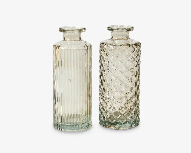 To klare glasflasker med brede, flade åbninger og cylindrisk form. Flasken til venstre har lodrette rillede linjer, der løber fra top til bund og giver en tekstureret overflade. Flasken til højre har et diamantmønstret, quiltet tekstur over hele overfladen. Begge flasker har tykke, solide bunde og et vintage-inspireret design.