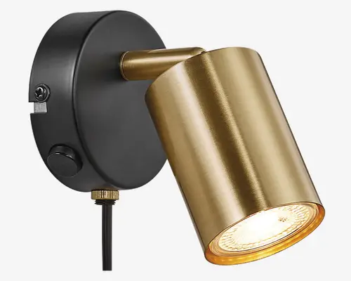 En vægmonteret spotlampe med et cylindrisk lampehoved i børstet messing og en mat sort, rund base. Lampehovedet kan justeres, så lyset kan rettes i forskellige retninger. Basen har en synlig drejeknap for nem betjening og en ledning, der går nedad, hvilket indikerer, at lampen er tilsluttet med stik. Designet er moderne og stilrent, velegnet til accent- eller arbejdsbelysning.