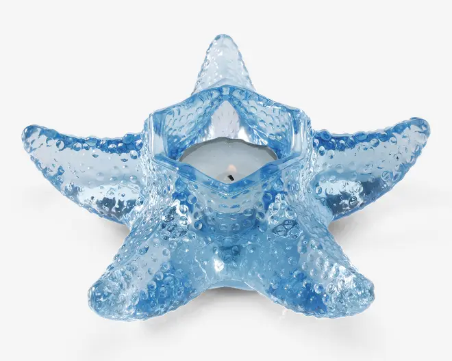 Et lysestage formet som en søstjerne, lavet af blå glas med en tekstureret overflade og små forhøjninger, der efterligner en rigtig søstjernes udseende. Holderen har et centralt hulrum, der passer til et lille fyrfadslys. Glasset er gennemsigtigt og giver et blødt, havinspireret udtryk.