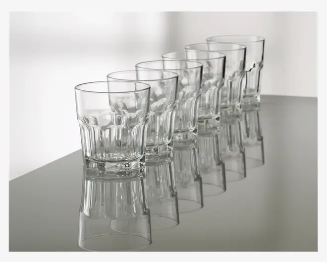 Sæt med seks klare glas tumblere arrangeret på række, hver med en facetteret nederste halvdel for bedre greb og en glat øverste kant. Glassene har en robust, tyk bund og en klassisk, let konisk form, velegnet til servering af vand, juice eller andre drikkevarer.