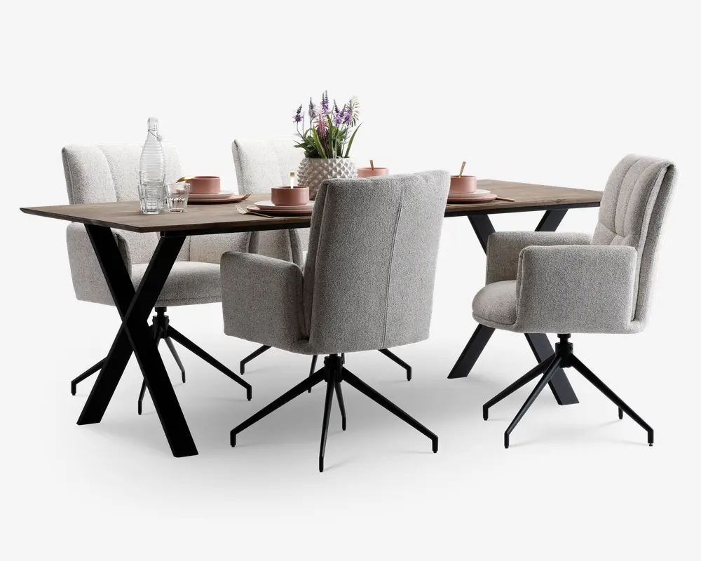 Moderne spisesæt med et rektangulært bord i træ og skrå sorte metalben. Sættet inkluderer fire polstrede stole i lysegråt stof, alle med armlæn og drejefod. Bordet er dækket med lyserøde keramiktallerkener, skåle og kopper samt klart glas og en dekorativ vase med lilla blomster.