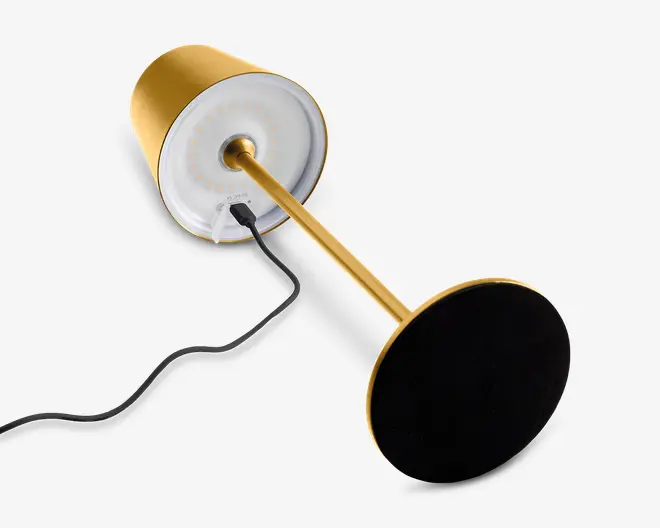 Elegant, moderne lampe med guldfarvet metalfinish, rund fod og cylindrisk lampeskærm. Undersiden af skærmen viser en ring af LED-lys samt en synlig USB-opladningsport med tilsluttet kabel. Lampen har et minimalistisk design med rene linjer og glat overflade.