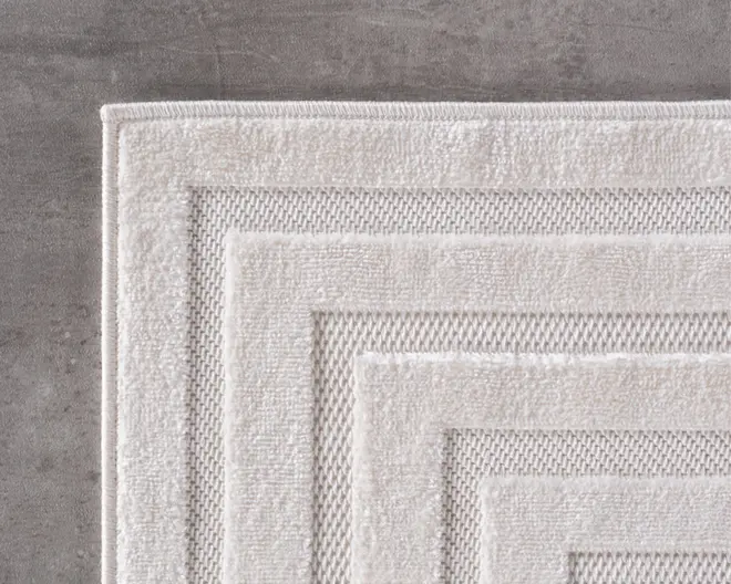 Nærbillede af en lysebeige rektangulær måtte med et tekstureret geometrisk mønster. Måtten har hævede kanter og skiftevis glatte og vævede teksturer, hvilket giver et lagdelt, moderne udseende. Kanterne er pænt afsluttede, og overfladen ser blød og fyldig ud.