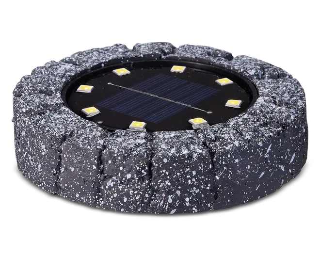 Rund udendørslampe med et robust, stenlignende ydre i mørkegrå med hvide pletter. I midten sidder et solpanel omgivet af seks jævnt fordelte LED-lys. Lampen er designet til at falde naturligt ind i have- eller sti-miljøer og giver solcelledrevet belysning.