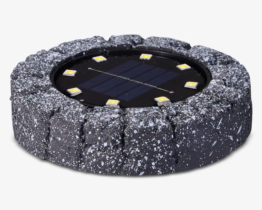 Rund udendørslampe med et robust, stenlignende ydre i mørkegrå med hvide pletter. I midten sidder et solpanel omgivet af seks jævnt fordelte LED-lys. Lampen er designet til at falde naturligt ind i have- eller sti-miljøer og giver solcelledrevet belysning.