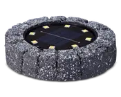 Rund udendørslampe med et robust, stenlignende ydre i mørkegrå med hvide pletter. I midten sidder et solpanel omgivet af seks jævnt fordelte LED-lys. Lampen er designet til at falde naturligt ind i have- eller sti-miljøer og giver solcelledrevet belysning.