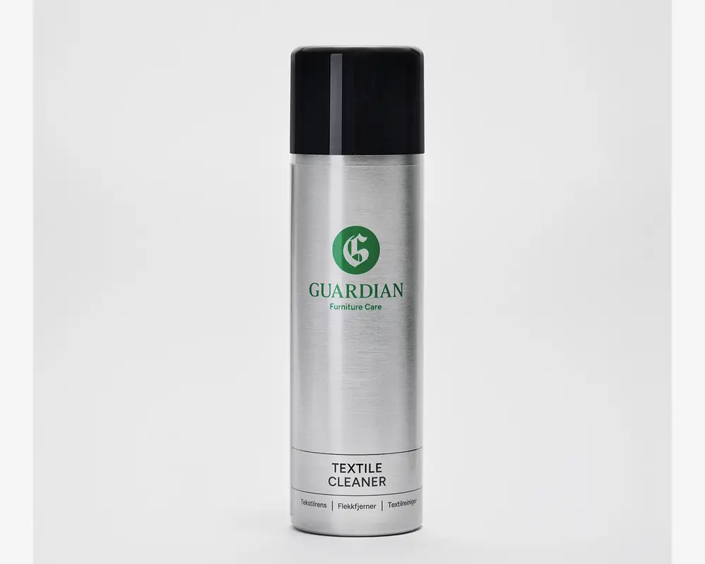 En høj, cylindrisk sølvfarvet spraydåse med sort låg. Dåsen har et grønt logo og mærket 'Guardian Furniture Care' trykt på forsiden. Nederst på etiketten står der 'Textile Cleaner' samt yderligere tekst på flere sprog. Designet er stilrent og moderne med en metallisk overflade.