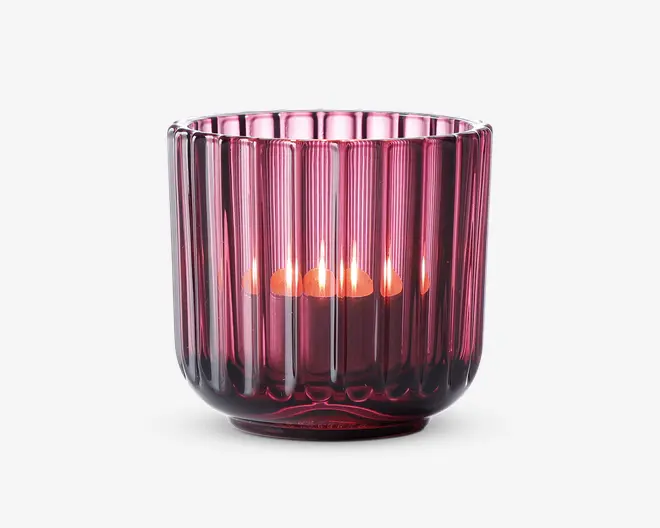 En dyb pink, rillet glasholder med lodrette riller, der indeholder et tændt lys. Glasset er gennemsigtigt, så den varme glød fra flammen skinner igennem og skaber en hyggelig stemning. Holderen har en rund form med let udadbuet kant og en tyk, solid bund.