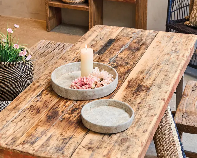 To runde, lave bakker lavet af lysegrå sten eller beton står på et rustikt træbord. Den største bakke indeholder et hvidt bloklys og nogle kunstige lyserøde blomster, mens den mindre bakke er tom. Bakkerne har et minimalistisk, naturligt udseende med glatte overflader og let hævede kanter.