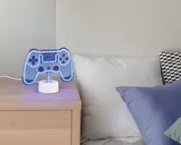 En bordlampe formet som en videospil-controller, oplyst med blåt LED-lys. Lampen har teksten 'GAME OVER' i midten og står på en rund, hvid base. Lampen er placeret på et natbord af lyst træ ved siden af en seng med hvide og blå puder.