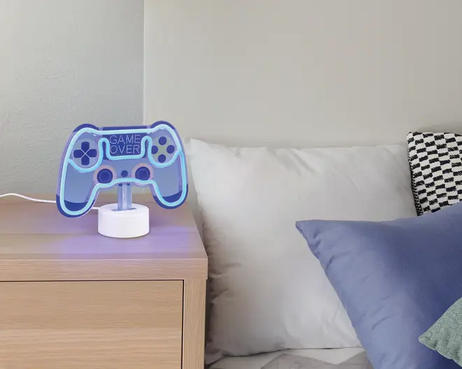 En bordlampe formet som en videospil-controller, oplyst med blåt LED-lys. Lampen har teksten 'GAME OVER' i midten og står på en rund, hvid base. Lampen er placeret på et natbord af lyst træ ved siden af en seng med hvide og blå puder.