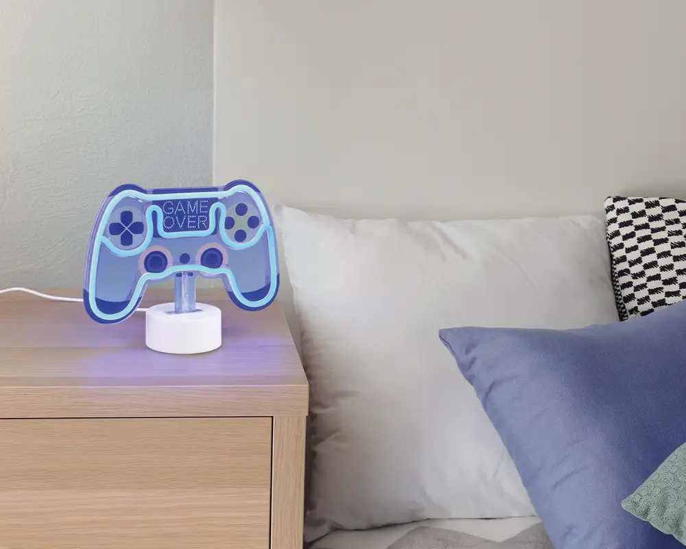 En bordlampe formet som en videospil-controller, oplyst med blåt LED-lys. Lampen har teksten 'GAME OVER' i midten og står på en rund, hvid base. Lampen er placeret på et natbord af lyst træ ved siden af en seng med hvide og blå puder.