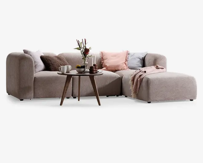 Moderne L-formet sofa betrukket med blødt, lysebeige stof, afrundede armlæn og lav ryg. Sofaen er pyntet med flere dekorative puder i forskellige teksturer og farver, herunder brun, rosa og lyseblå. Et hyggeligt tæppe er lagt over chaiselongen. Foran sofaen står et lille rundt sofabord i træ med en vase med blomster, to stearinlys, en kop med underkop og en lille skål, hvilket giver et varmt og indbydende udtryk.