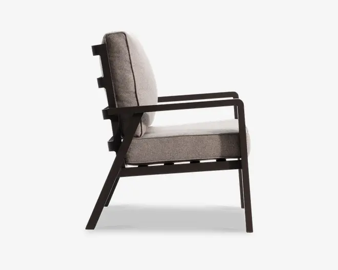 Stol set fra siden med en tyk, polstret sæde- og ryghynde betrukket med tekstil. Stellet er lavet af mørkt træ med skrå ben og armlæn, hvilket giver et moderne og minimalistisk udtryk. Armlænene er flade og går lige fra ryggen til forsiden, hvilket giver god støtte.