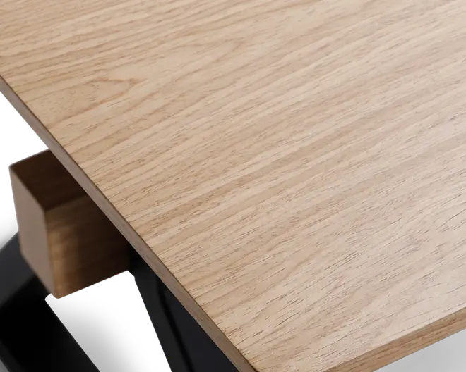 Nærbillede af en bordplade i lyst egetræ med en glat, mat overflade. Bordpladen har et naturligt træmønster og let afrundede kanter. Bordets hjørne understøttes af en solid sort metalramme med en trædetalje, hvilket giver et moderne og minimalistisk udtryk.