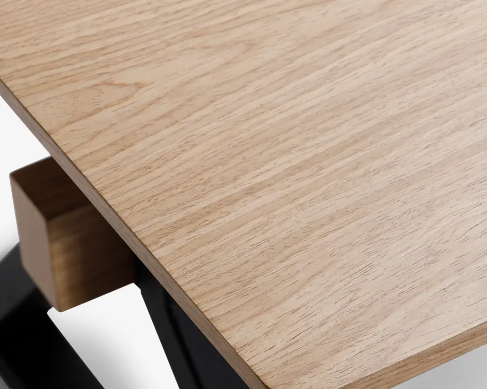 Nærbillede af en bordplade i lyst egetræ med en glat, mat overflade. Bordpladen har et naturligt træmønster og let afrundede kanter. Bordets hjørne understøttes af en solid sort metalramme med en trædetalje, hvilket giver et moderne og minimalistisk udtryk.
