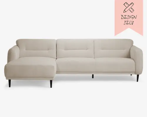 Moderne hjørnesofa i en lys beige farve med chaiselong til venstre. Betrækket er i et blødt, tekstureret stof. Sofaen har rene linjer, diskrete knapdetaljer på ryglænene og slanke, sorte, koniske ben. Armlænene er let afrundede, og det samlede design er minimalistisk og moderne, hvilket passer til mange forskellige stuer.