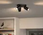 Sort loftspot med tre lyskilder, rund base og justerbare cylindriske lampehoveder, vist tændt mod et lysegråt loft.