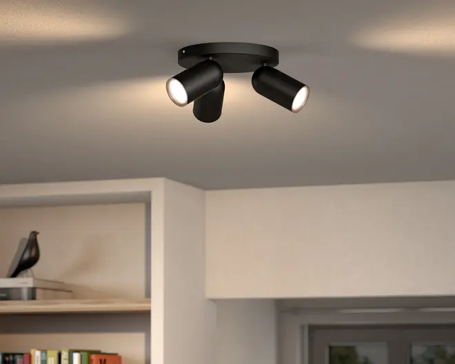 Sort loftspot med tre lyskilder, rund base og justerbare cylindriske lampehoveder, vist tændt mod et lysegråt loft.