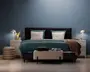 Et moderne soveværelse med en seng, der har et mørkt polstret hovedgærde, teal sengetøj og to store, bløde hvide puder. Sengen er flankeret af to hvide natborde, hver med en moderne lampe og dekorative genstande som en vase med gule blomster og en bakke med en karaffel. For enden af sengen står en tekstureret, lysefarvet bænk med sammenfoldede tæpper og en pude ovenpå. Rummet har et minimalistisk design med en blå accentvæg og lyst trægulv.